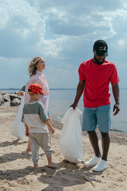 A person standing outdoors on a grassy area, wearing a checked shirt, green gloves, and beige trousers, holding open a large black rubbish bag made of plastic. The bag's top rim is folded over, reveal