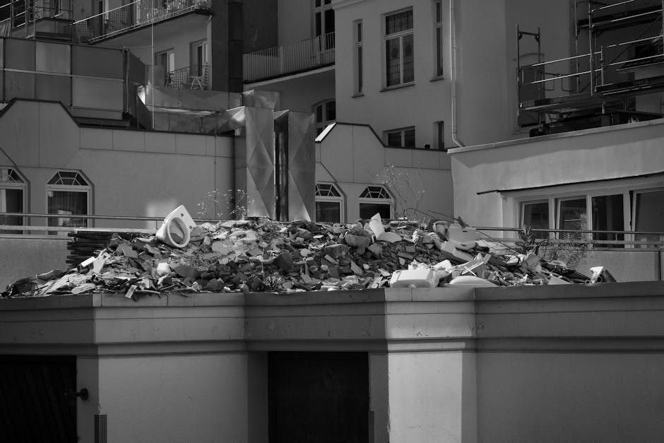 A pile of demolished building debris situated on a paved area, consisting of various materials including broken bricks, concrete chunks, wooden fragments, and pieces of blue tarpaulin draped over the 