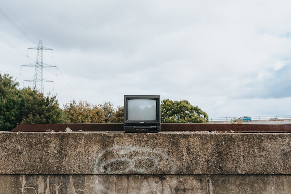 A collection of old, discarded television sets of varying sizes and styles, primarily with built-in cathode ray tube screens and wooden or plastic casings, are piled haphazardly on a sandy outdoor sur